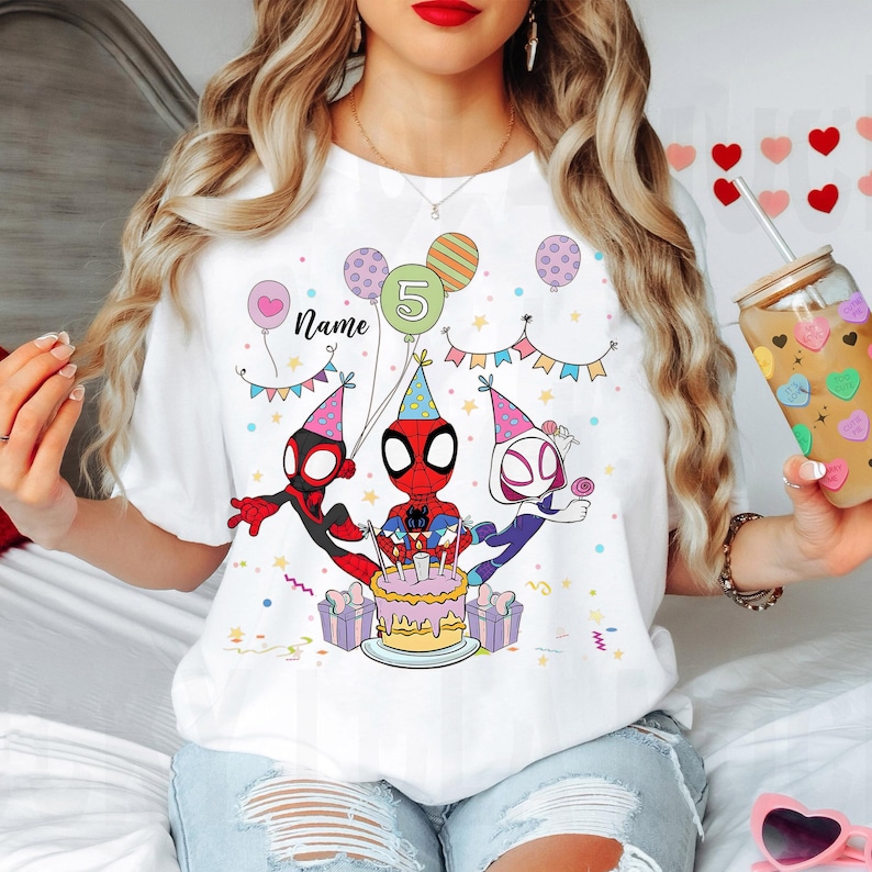 May include: White t-shirt featuring a colorful graphic of Spider-Man characters celebrating a birthday. The design includes balloons, a cake, and the number 5. The word "Name" is also included in the design.
