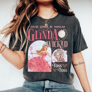 May include: Dark gray t-shirt featuring the text "You're Gonna Be Popular" and "Glinda Wicked". The graphic includes an illustration of a woman in a pink dress, a crown, and pink shoes. The shirt has a vintage look.