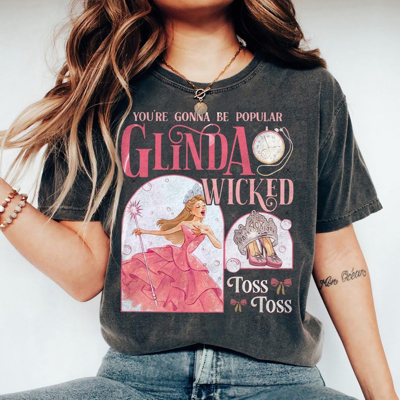 May include: Dark gray t-shirt featuring the text "You're Gonna Be Popular" and "Glinda Wicked". The graphic includes an illustration of a woman in a pink dress, a crown, and pink shoes. The shirt has a vintage look.