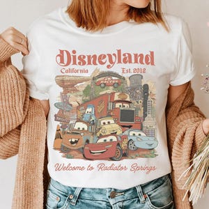 May include: White t-shirt featuring a colorful graphic of characters from the movie Cars, with the text "Disneyland California Est. 2012" and "Welcome to Radiator Springs". The design includes various animated cars and a desert landscape.