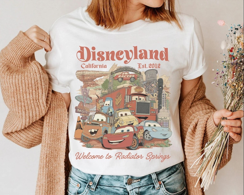 May include: White t-shirt featuring a colorful graphic of characters from the movie Cars, with the text "Disneyland California Est. 2012" and "Welcome to Radiator Springs". The design includes various animated cars and a desert landscape.