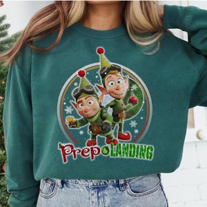 Könnte beinhalten: Türkisgrünes Sweatshirt mit einem kreisförmigen Aufdruck von zwei Cartoon-Elfen in grünen Outfits und Schutzbrillen. Die Elfen sind vor dem Hintergrund von Schneeflocken und einem Regenbogen platziert. Der Text "Prep & Landing" steht darunter.