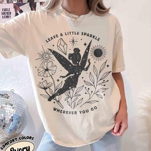 May include: Ivory-colored t-shirt with a black graphic of a fairy, flowers, and the text "LEAVE A LITTLE SPARKLE WHEREVER YOU GO." The shirt also has the text "I SHOULD HAVE BEEN A COWBOY" on the upper left.