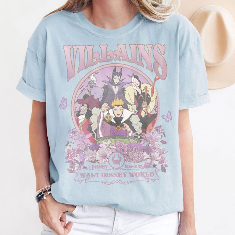 May include: Light blue t-shirt featuring a graphic of Disney villains. The design includes the word "Villains" in a pink, retro font, surrounded by illustrations of various characters and floral elements. The text "Disney Villains Walt Disney World" is also present.