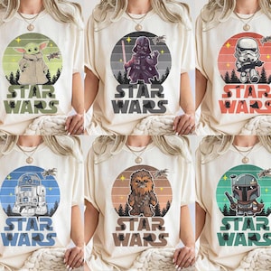 Pode incluir: T-shirts creme com personagens de Star Wars como Baby Yoda, Darth Vader e um Stormtrooper. Cada design inclui um gr&aacute;fico circular com o personagem, uma cena de floresta e as palavras "STAR WARS" em negrito.