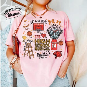 May include: A light pink t-shirt with a graphic design featuring basketballs, a basketball hoop, and the text "EHS High School Musical." The shirt also includes the text "Get'cha Head in the Game" and "Breaking Free."