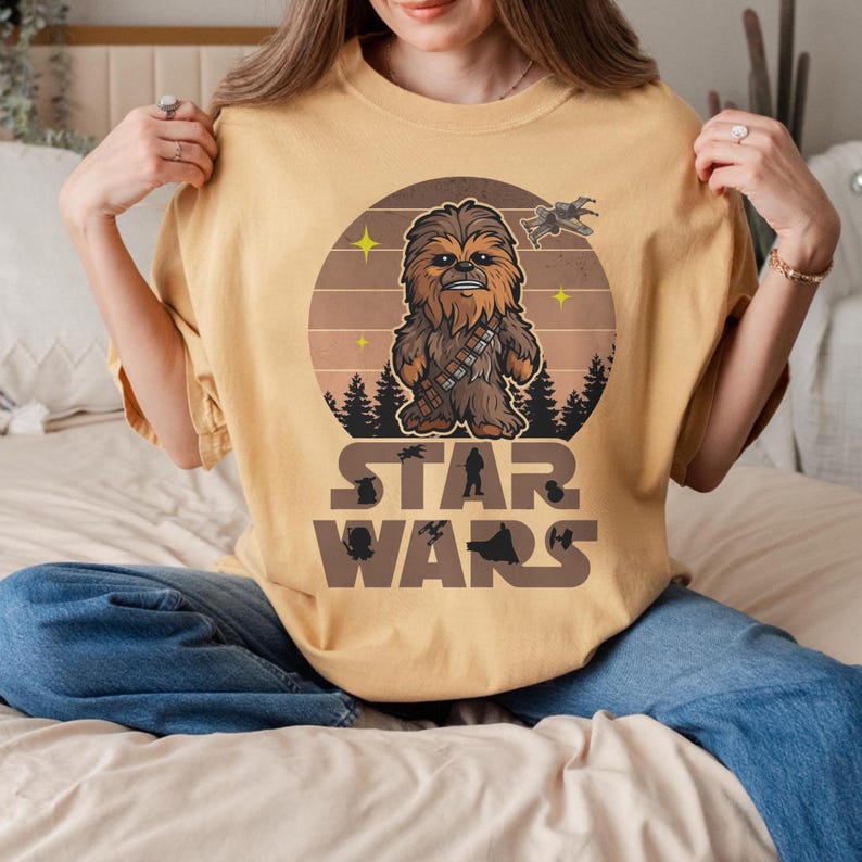 Pode incluir: Camiseta amarelo mostarda com um desenho circular de Chewbacca em tons de castanho e bege. O logotipo de Star Wars &eacute; impresso em castanho por baixo. A camisa tem um corte folgado.
