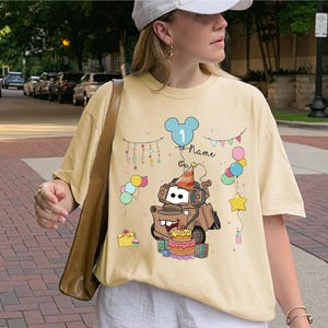 May include: A light yellow t-shirt featuring a cartoon image of Mater from the movie Cars, wearing a party hat and surrounded by balloons and a birthday cake. The shirt has the number "1" and the word "Name" on it.