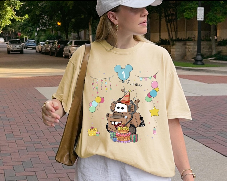 May include: A light yellow t-shirt featuring a cartoon image of Mater from the movie Cars, wearing a party hat and surrounded by balloons and a birthday cake. The shirt has the number "1" and the word "Name" on it.