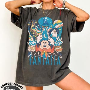 May include: Dark gray t-shirt featuring a colorful graphic of Mickey Mouse in a wizard hat with the word "Fantasia" in blue. The design includes elements from the Disney film, such as the Sorcerer's hat and musical notes. The shirt is from Comfort Colors.
