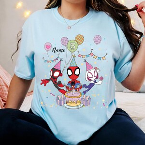 May include: Light blue t-shirt featuring a birthday design with Spider-Man characters. The design includes a cake, balloons, and the number 5. The word "Name" is also present. The shirt is decorated with colorful confetti and gift boxes.