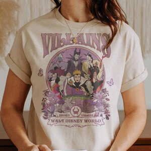 May include: Beige t-shirt featuring a purple graphic of Disney villains, including Maleficent and the Evil Queen, surrounded by flowers. The word "VILLAINS" is arched above the characters, with "WALT DISNEY WORLD" below.