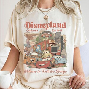 May include: Cream-colored t-shirt featuring a graphic of characters from the movie Cars, with the text "Disneyland California Est. 2012" and "Welcome to Radiator Springs". The design includes various cartoon cars and a desert landscape.