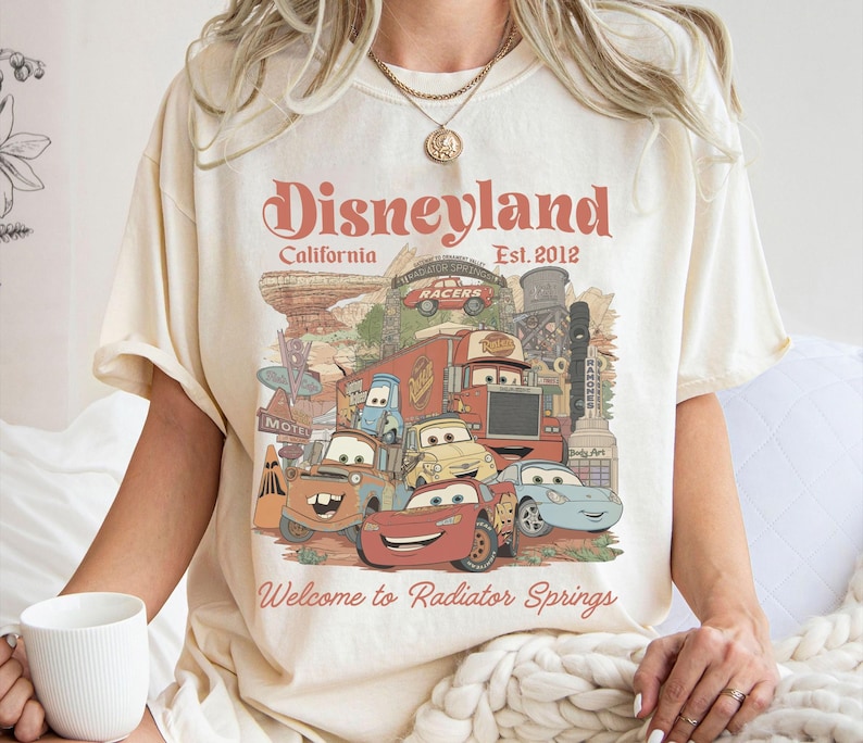 May include: Cream-colored t-shirt featuring a graphic of characters from the movie Cars, with the text "Disneyland California Est. 2012" and "Welcome to Radiator Springs". The design includes various cartoon cars and a desert landscape.