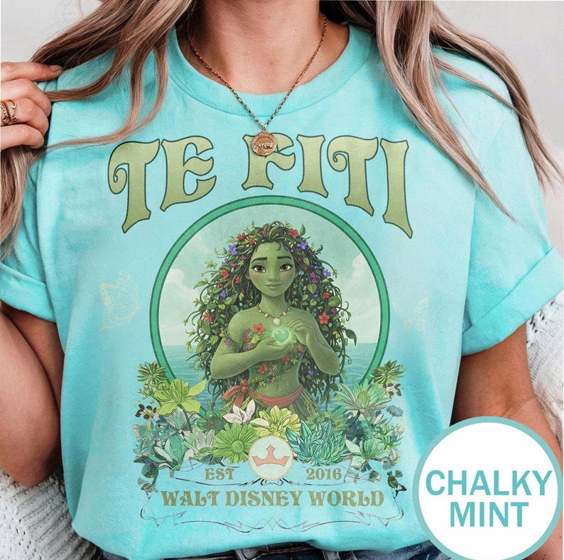 May include: A light blue t-shirt featuring the text "TE FITI" above a circular graphic of a woman with green skin and floral adornments. The graphic includes the text "EST 2016 WALT DISNEY WORLD". The shirt has short sleeves.