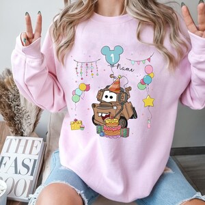 May include: A light pink sweatshirt featuring a cartoon illustration of a brown tow truck with a birthday cake and party hat. The design includes balloons, a star, and the number "1" inside a Mickey Mouse head. The word "Name" is also included.