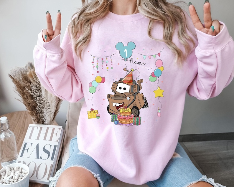 May include: A light pink sweatshirt featuring a cartoon illustration of a brown tow truck with a birthday cake and party hat. The design includes balloons, a star, and the number "1" inside a Mickey Mouse head. The word "Name" is also included.