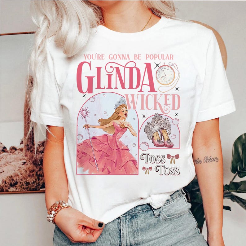 May include: White t-shirt featuring a pink graphic with the text "You're Gonna Be Popular Glinda Wicked." The graphic includes an illustration of a woman in a pink dress, a crown, and a clock. The shirt has a crew neck and short sleeves.