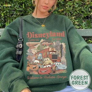 May include: Forest green sweatshirt featuring a graphic of Disneyland California and the text "Welcome to Radiator Springs". The graphic includes cartoon cars and a truck. The sweatshirt has a crew neck.