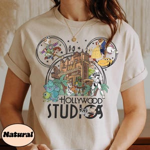 May include: Beige t-shirt featuring a colorful graphic of Disney characters and the words "Hollywood Studios." The design includes characters from Toy Story, Beauty and the Beast, and Star Wars. The shirt has short sleeves and a round neckline.