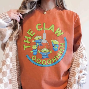 Può includere: T-shirt arancione con una grafica degli alieni del film Toy Story. Il design include il testo "THE CLAW" in verde, con gli alieni in uniformi blu e il testo "OOOOOHHH" in blu.