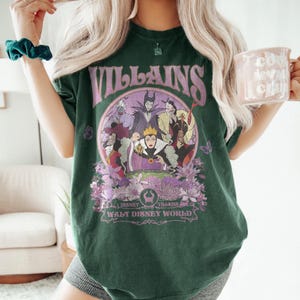 May include: Dark green t-shirt featuring a graphic of Disney villains in a circular design with the word "VILLAINS" above. The design includes the text "Disney Villains Walt Disney World" below the image. The shirt has a relaxed fit.