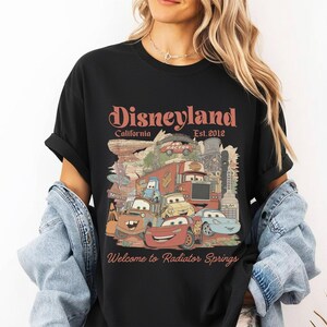 May include: Black t-shirt featuring a graphic of Disney Cars characters and the text "Disneyland California Est. 2012" and "Welcome to Radiator Springs". The design includes various cartoon cars and buildings.