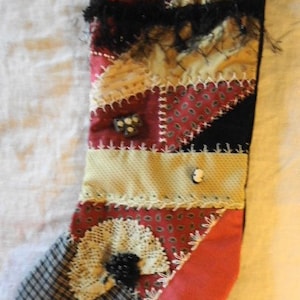 May include: Handmade Christmas stocking with a patchwork design. The stocking features various fabrics in red, black, cream, and pink, with decorative stitching and embellishments. A black loop is attached for hanging.
