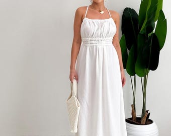 Op de afbeelding: Een witte maxi-jurk met een halterhalslijn en een geplooid lijfje. De jurk heeft een getailleerde taille en een zwierige rok. Een bijpassende witte handtas wordt vastgehouden. Een grote groene plant staat op de achtergrond.