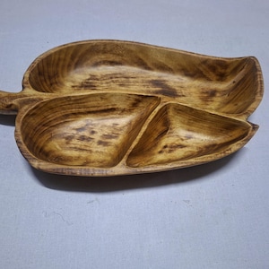Puede incluir: Bandeja de madera con forma de hoja con tres compartimentos. La bandeja está hecha de madera de color marrón claro con vetas de madera más oscuras. Tiene un asa en un extremo y está diseñada para servir aperitivos o entremeses.
