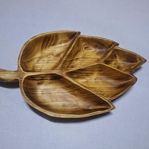 Puede incluir: Una bandeja de madera con forma de hoja con múltiples compartimentos. La bandeja está hecha de madera de color marrón claro con vetas de color marrón más oscuro. Tiene un asa en un lado y está diseñada para servir aperitivos.