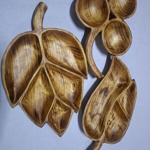 Puede incluir: Tres fuentes de madera para servir, con forma de hojas y un conjunto de cuencos. Las fuentes en forma de hoja tienen múltiples compartimentos. La madera tiene un tono cálido y natural con vetas visibles.