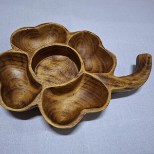 Puede incluir: Una bandeja de madera para servir con forma de trébol. Tiene cinco compartimentos y un asa. La madera es de color marrón cálido con vetas visibles. Esta bandeja se utiliza probablemente para servir aperitivos, y se mide en centímetros.