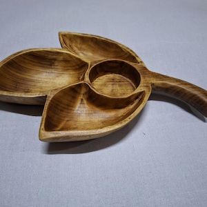 Puede incluir: Bandeja de madera para servir con forma de hoja, con tres compartimentos y un cuenco central, más un asa. La madera tiene un tono cálido y natural, mostrando la veta. Ideal para servir aperitivos o pequeños platos.