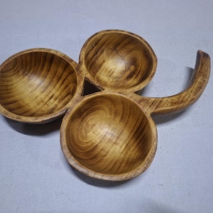 Puede incluir: Un plato de madera para servir con tres cuencos conectados y un asa curva. Los cuencos son circulares con un acabado marrón claro y veta de madera visible. El asa se extiende desde el cuenco superior.