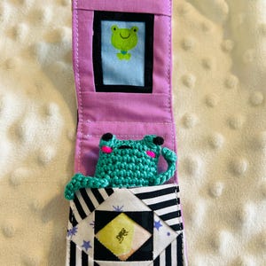 May include: A handmade, open-faced fabric case with a frog theme. The case is pink with a black and white striped pocket. A crocheted green frog is nestled in the pocket. A small picture of a frog is in a frame at the top.