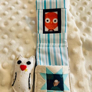 May include: A handmade fabric owl toy and a soft book with an owl theme. The book features a striped cover with an orange owl illustration and a patchwork design. The owl toy is white with black and red embroidered details.