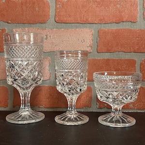 May include: Three clear, vintage-style crystal glasses with diamond and crosshatch patterns. The glasses vary in size, with a tall goblet, a smaller stemmed glass, and a low-profile dessert dish. The glasses are displayed against a brick wall.