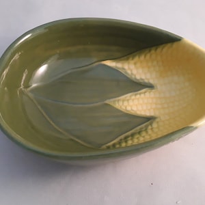 May include: A green and yellow ceramic bowl shaped like an ear of corn. The bowl has a textured surface and is designed to hold corn on the cob.