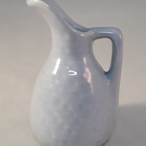 May include: A small, light blue ceramic pitcher with a rounded body and a curved spout. The pitcher has a white glaze with a subtle marbled effect.