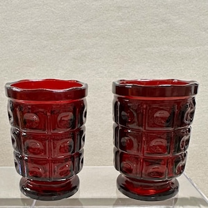 May include: Two ruby red vintage drinking glasses. Each glass has a cylindrical shape with a textured pattern of squares and circles. The rims have a wavy design. The glasses are made of glass and are displayed on a clear surface.