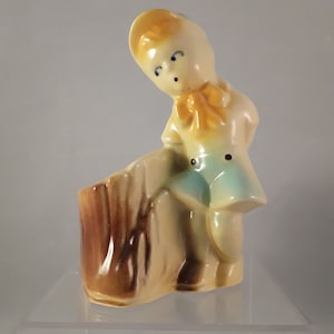 May include: A ceramic planter shaped like a boy wearing a yellow hat and blue pants. The planter is standing on a tree stump.