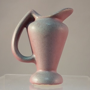 May include: A small, pink and blue ceramic pitcher with a textured glaze. The pitcher has a wide, rounded spout and a curved handle.