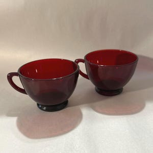 May include: Two ruby red glass teacups with handles. The cups have a rounded bowl shape and sit on a black base. The cups are made of a transparent glass material.