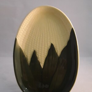 May include: A ceramic serving dish shaped like an ear of corn. The dish is yellow with a green and brown design.