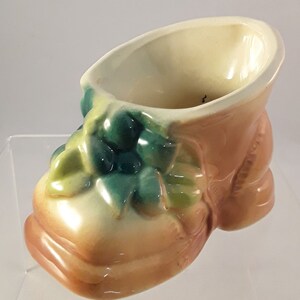 May include: A ceramic planter shaped like a brown shoe with green leaves. The planter is a vintage style and has a cream colored interior.
