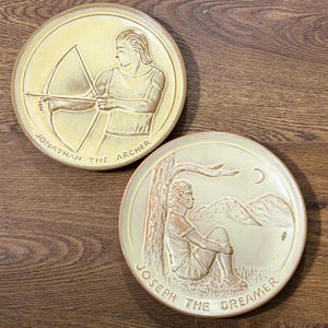 May include: Two round, cream-colored ceramic plates with raised relief designs. One plate features an archer with the text "Jonathan the Archer." The other plate depicts a seated figure with the text "Joseph the Dreamer."