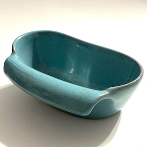 May include: A turquoise ceramic dish with a curved handle on one side. The dish has a rectangular shape with rounded edges and a textured surface. The rim has a darker, brownish hue.
