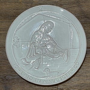 May include: A round, off-white ceramic plate with a relief design depicting a religious scene. The image features a woman, a cow, and a lamb, with the text "19 - LAID IN A MANGER - 69" around the edge. The plate has a raised border.