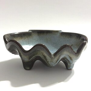 May include: A ceramic bowl with a wavy rim and three small feet. The bowl has a blue-green interior and a dark brown rim. The exterior has a mottled, glazed finish. The bowl is likely handmade and suitable for serving or display.